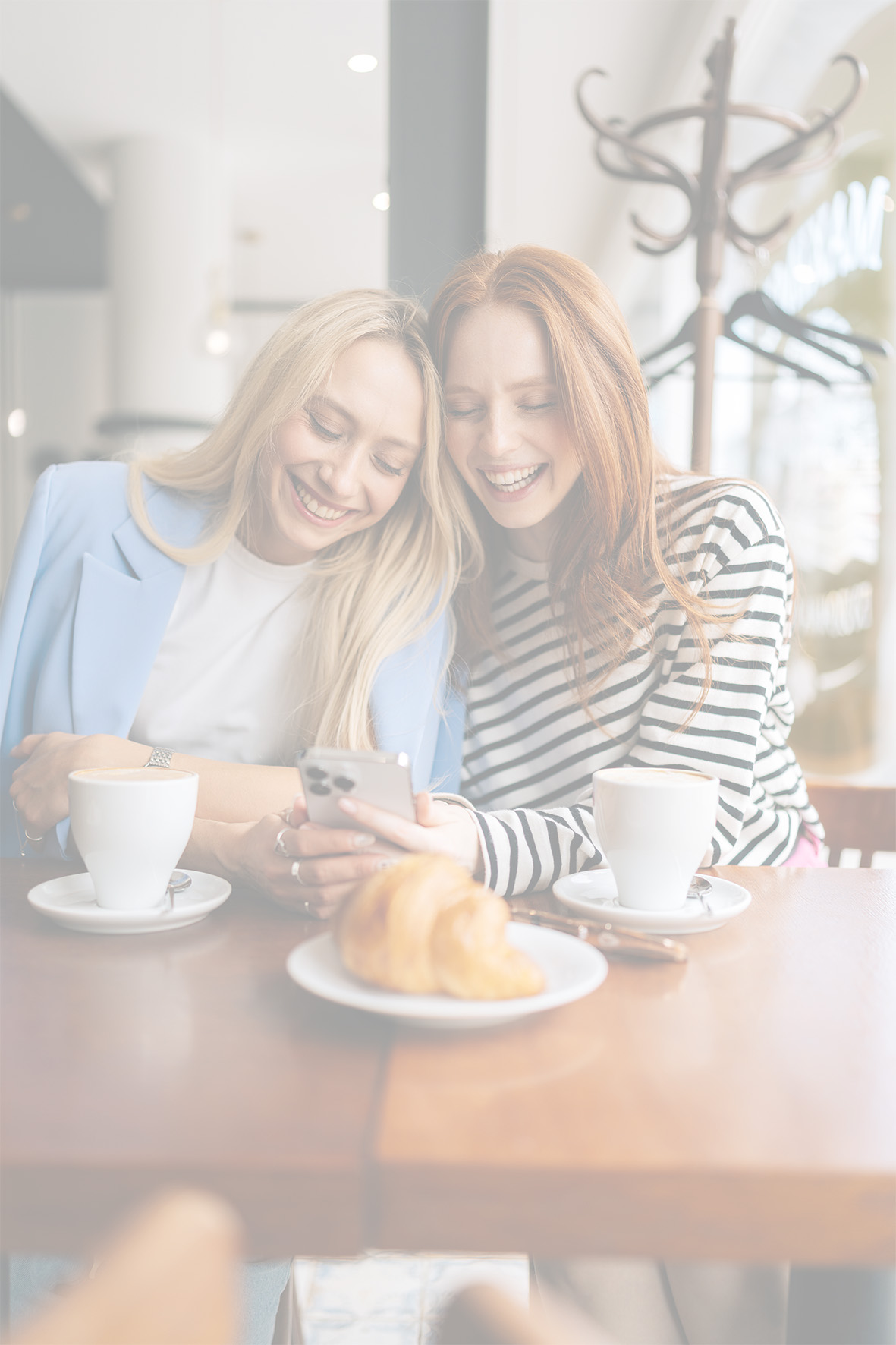 Zwei Frauen lachen gemeinsam an einem Tisch in einem Café. Vor ihnen stehen Kaffeetassen und ein Croissant